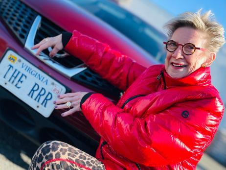 Gabie wearing a red coat standing in front of a red car, pointing at the license plate.