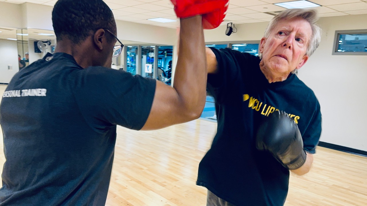 Walter Dotts practices light sparring in a gym studio, wearing boxing gloves and a dark T‑shirt, as a trainer holds up a focus mitt to block and guide a punch; wood floors, wall mirrors, and fitness equipment appear in the background.