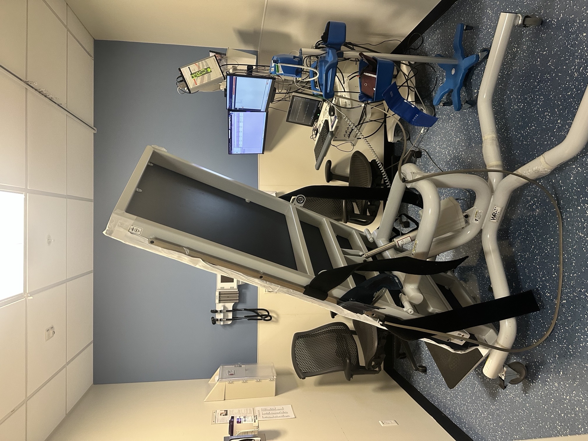 **ALT text (WCAG-compliant):**   Tilt table in a clinical exam room, positioned upright at an angle, with padded surface, safety straps, footboard, and adjustable supports, connected to nearby monitors and medical equipment for patient testing.